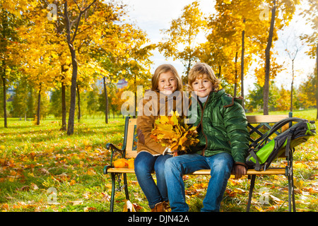 Couple happy blond kids sitting on the bench in the autumn park with bouquet of maple leaves Stock Photo