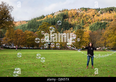 A woman makes lots of small bubbles with sticks and string loops on village green in Betws-y-Coed, Conwy, North Wales, UK Stock Photo