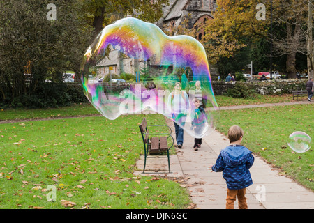 A young boy has fun with a giant soap bubble showing rainbow colours and people smiling in Betws-y-Coed, Conwy, North Wales, UK Stock Photo