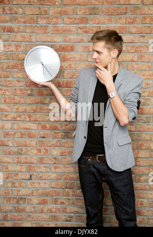 Young businessman holding a clock on brick wall background. Time in business concept Stock Photo