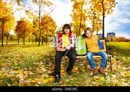 Two happy boys sitting on the bench with backpack in autumn park Stock Photo