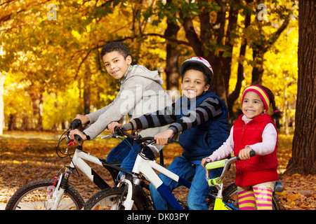 Close portrait of three black boys and girl, brothers and sister riding bicycles and scooter in the autumn October park with maple leaves Stock Photo