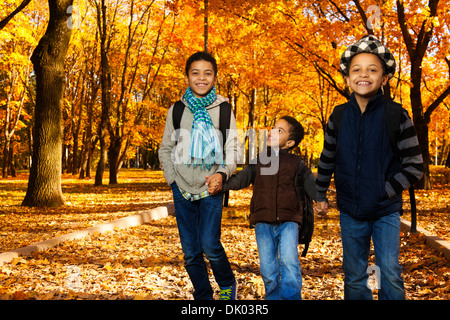 Three black boys, happy brothers 5-10 years old going together holding hands in the park wearing backpacks and autumn clothes in maple park Stock Photo