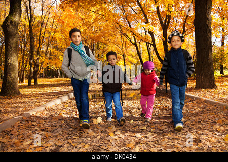 Group of four black boys and girl, happy brothers and sister 3-10 years old going together holding hands in the park wearing backpacks and autumn clothes in oak park with orange leaves Stock Photo