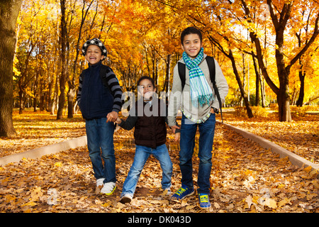 Three happy, laughing black boys, happy brothers 5-10 years old going together holding hands in the park wearing backpacks and autumn clothes in maple park Stock Photo