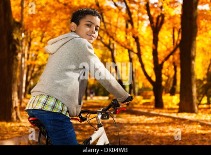 Close portrait of nice black boy riding a bicycle in the autumn park full of autumn leaves turning back Stock Photo