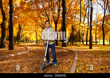 One handsome black 10 years old boy riding a scooter in autumn park with maple orange leaves wearing autumn sweatshirt with hood Stock Photo