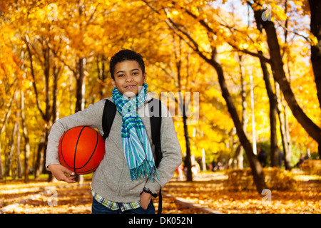 Handsome black boy 10 years old standing in the autumn park under maple trees with orange basketball ball wearing casual clothes with scarf and sweatshirt Stock Photo