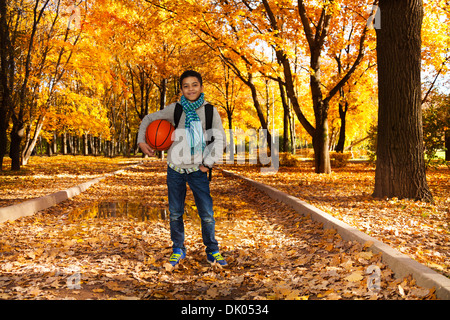 Handsome smiling black boy 10 years old standing in the autumn park under maple trees with orange basketball ball wearing casual clothes with scarf and sweatshirt Stock Photo