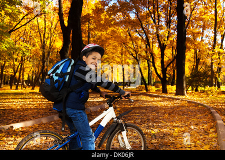 Happy smiling 8 years old black boy with backpack riding a bike in the autumn park full of orange leaves leaning on bicycle stern turning back Stock Photo