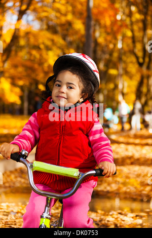 Close portrait of a little black 3 years old black girl riding a bicycle in the park holding stern and wearing helmet Stock Photo