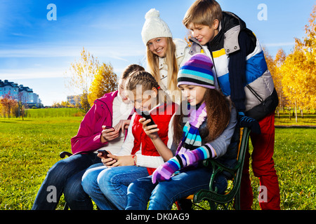 Group of five school age teen kids busy looking at their mobile phones sitting on the bench in autumn park on sunny day Stock Photo