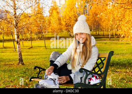 Smiling blond teen girl with long hair sitting on the bench in the park and putting on roller blades Stock Photo
