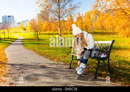 Blond teen girl with long hair in autumn park sitting on the bench and putting on roller blades Stock Photo