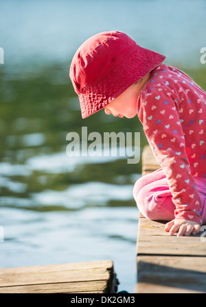 Lifestyle moment of summer childhood. Little girl sitting on jetty, looking down. Stock Photo