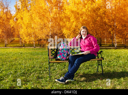 Beautiful teen 14 years old school girl sitting on the bench in autumn park with backpack and textbooks Stock Photo