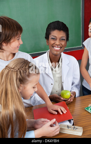 Teacher Holding Apple With Students Standing At Desk Stock Photo