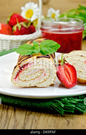 Biscuit roulade with cream and jam, a jar of jam, napkin, chamomile flowers, strawberries, mint on the wooden boards Stock Photo