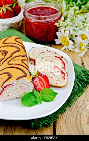 Biscuit roulade with cream and jam, a jar of jam, napkin, chamomile flowers, strawberries, mint on a wooden board Stock Photo
