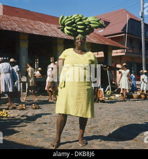 Dominica, Caribbean, Roseau market woman carrying a large stalk of green bananas on her head. Stock Photo