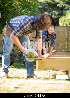 Carpenter Assisting Coworker In Cutting Wood With Handheld Saw Stock Photo