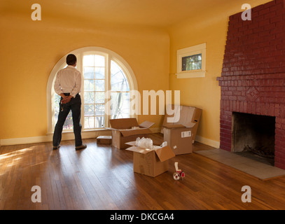 A man looks out of an empty room in a residential house and cardboard boxes suggesting he is moving in or out. Stock Photo