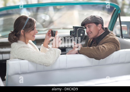 Couple in convertible filming and photographing each other with modern and vintage equipment Stock Photo