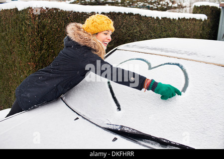 Woman drawing heart shape on snow covered windscreen Stock Photo