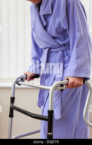 Elderly senior woman wearing a purple bathrobe holding using a zimmer frame walker or support trolley for walking in a home. England UK Britain Stock Photo