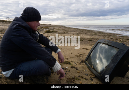 Man watching a television set washed up on a beach. Stock Photo