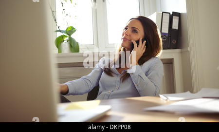 Attractive young woman sitting at her desk talking on mobile phone. Pretty caucasian businesswoman working in home office. Stock Photo