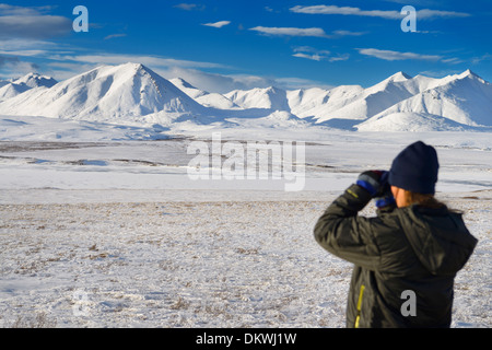 Female tourist looking at the snow covered Brooks Range mountains with binoculars from the isolated Dalton Highway Alaska USA Stock Photo