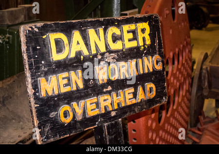 Sign warning of potential danger with men working overhead in a museum devoted to railway engineering Stock Photo