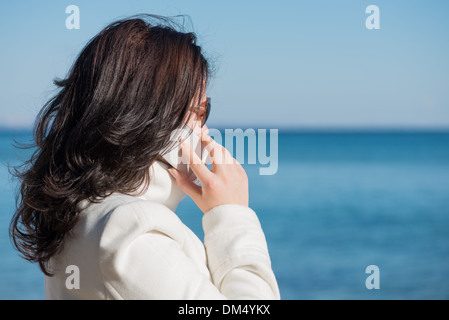 Young woman is speaking by mobile phone at the seaside and looking at the sea. It's spring time. Stock Photo