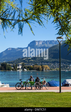 Couple with bicycles sitting on bench looking at the view over Lake Annecy, Savoie, France Stock Photo