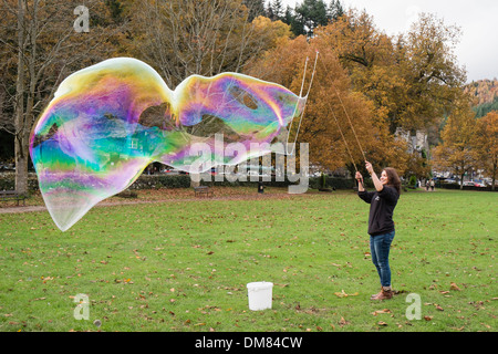 A woman makes a giant soap bubble with rainbow colours using a bubble wand in Betws-y-Coed, Conwy, North Wales, UK, Britain Stock Photo