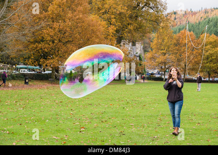 A woman makes a giant soap bubble with rainbow colours using a bubble wand in Betws-y-Coed, Conwy, North Wales, UK, Britain Stock Photo