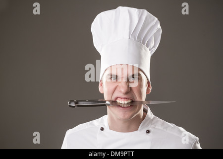 Portrait of smiling chef with knife in mouth isolated on grey background Stock Photo