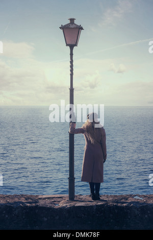 a woman in a pink coat is standing next to a lantern at the sea Stock Photo