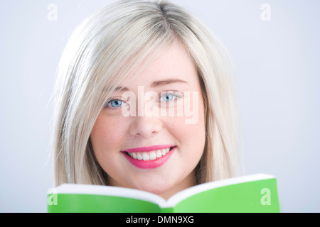 Pretty blonde teenager peeking over a green book held in front of her face. Stock Photo