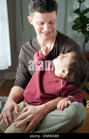 A child sits in his father's lap, pointing his finger at his father who is smiling. A family portrait. Stock Photo