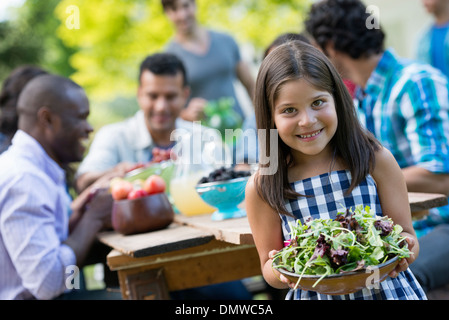 Adults and children around a table in a garden. A child holding a bowl of salad. Stock Photo