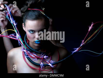 A robot like woman staring at camera with sparkling electric cables wrapped around her neck. Hands are adjusting cables. Stock Photo