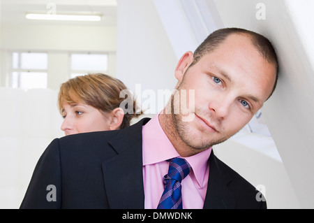 A young man in a suit and pink shirt leaning against a wall and a young woman in  background. Stock Photo