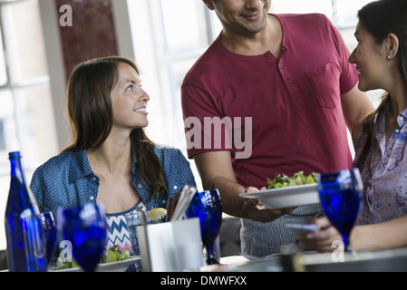 A cafe interior. A man in a waiter's apron serving a meal to two women at a table. Stock Photo