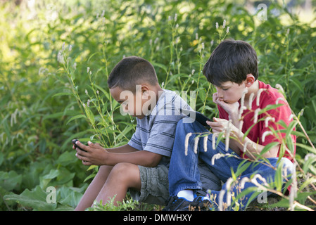 Two boys sitting in a field one on a smart phone and one using a digital tablet. Stock Photo
