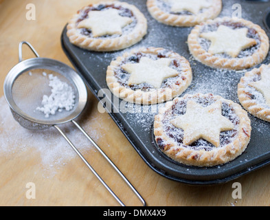 Home made icing sugar dusted mince pies in baking tray with sieve on wooden surface Stock Photo