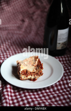 A slice of homemade meat lasagna on a white plate with a bottle of red wine in the background Stock Photo