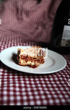 A slice of homemade meat lasagna on a white plate with a bottle of red wine in the background Stock Photo