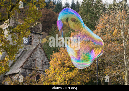 A giant soap bubble showing rainbow colours floats in air towards church clock tower in Betws-y-Coed, North Wales, UK, Britain Stock Photo